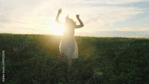 Girl child in a dress runs across the grass to meet the setting sun and waves her arms like a bird flapping her wings. Slow motion. Concept of childhood dreams, freedom, unity with nature. 