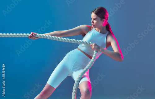 Fototapeta Naklejka Na Ścianę i Meble -  A sporty girl with brown hair pulls the rope. Isolated athletic figure of a beautiful girl in a white uniform on a blue background.