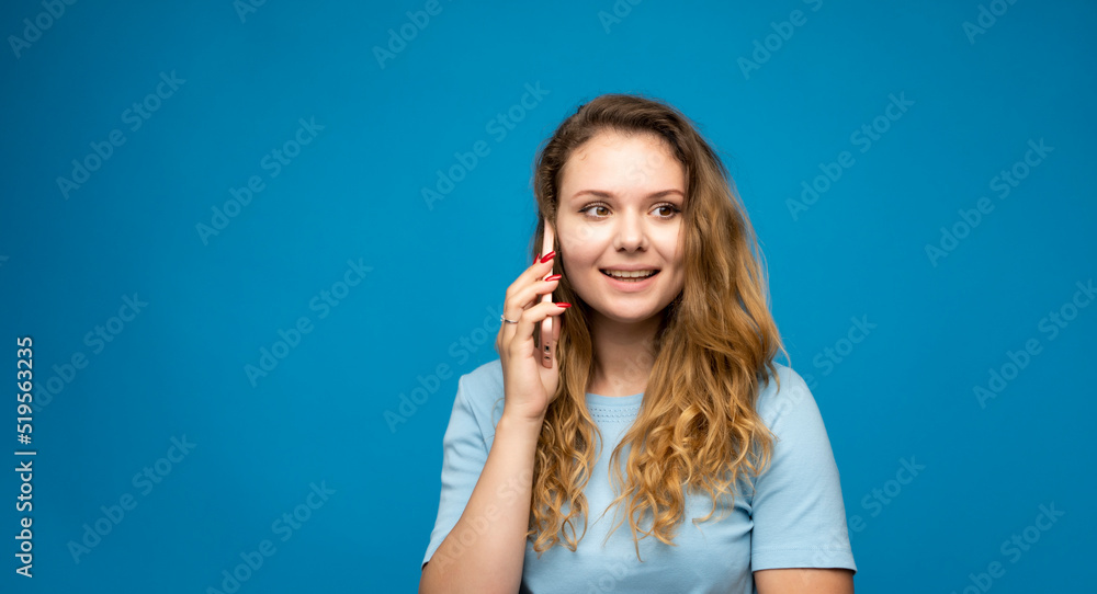Portrait of a surprised girl with a curly hair dressed in blue t-shirt talking on mobile phone isolated over blue background. Portrait of a surprised girl with a curly hair dressed in blue t-shirt talking on mobile phone isolated over blue background.
