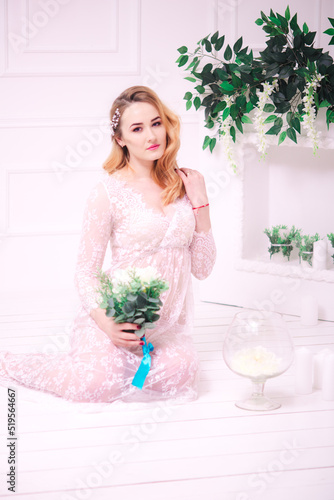 Young attractive bride with blond hair with wedding makeup and hairstyle in a white lace peignoir with bride's bouquet in a bright interior