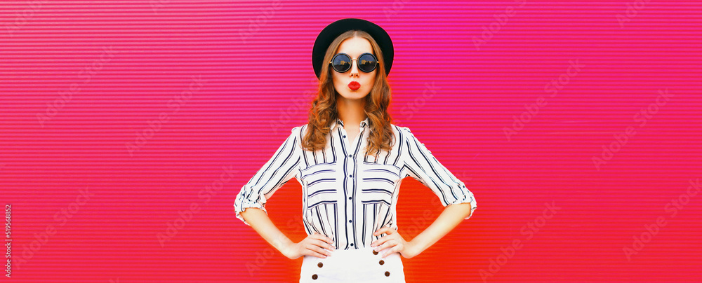 Fototapeta premium Portrait of beautiful young woman blowing her lips sending air kiss wearing sunglasses, summer straw hat, striped shirt on pink background