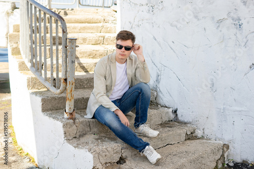 Young man with sunglasses in stylish clothes sits on steps outside