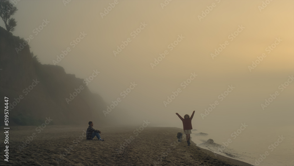 Couple enjoying sunrise on beach. Man sitting on shore. Woman walking along sea