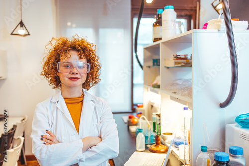 Young female scientist working at the laboratory. Close-up of serious laboratory worker holding ampoule in front of eyes and examines contents. Scientist in protection gloves eyewear and white coat. 