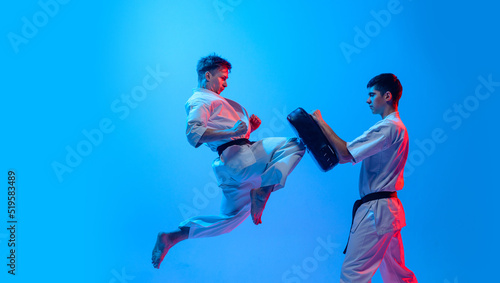 Fototapeta Naklejka Na Ścianę i Meble -  Studio shot of sports training of two karatedo fighters in doboks isolated on blue background in neon. Concept of combat sport, challenges, skills