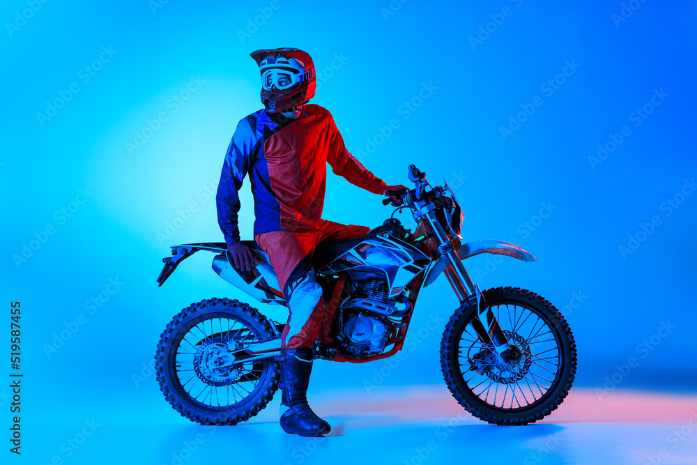 Portrait of young man, biker in professional sports equipment isolated over blue studio background in neon light. Resting Portrait of young man, biker in professional sports equipment isolated over blue studio background in neon light. Resting