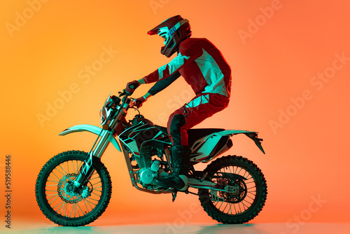 Portrait of young man, biker in full equipments riding motorbike isolated over orange studio background in neon light. Doing tricks