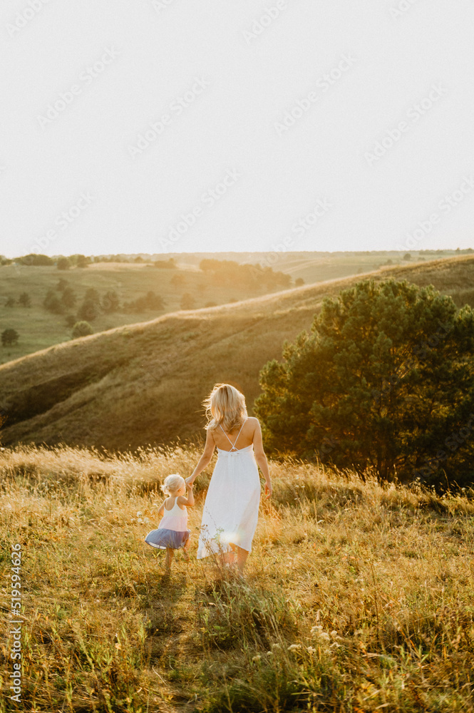 A beautiful young mother with a little daughter are hugging and walking across the field at sunset.