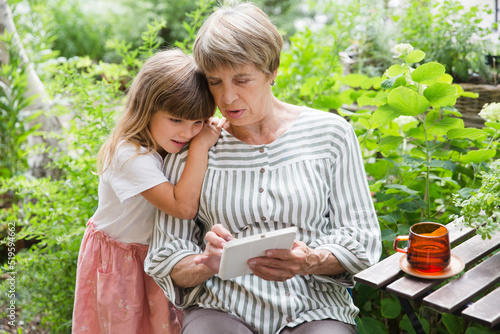 grandmother and little granddaughte using tablet and having fun together in the garden. Happy family enjoying summer outdoors.