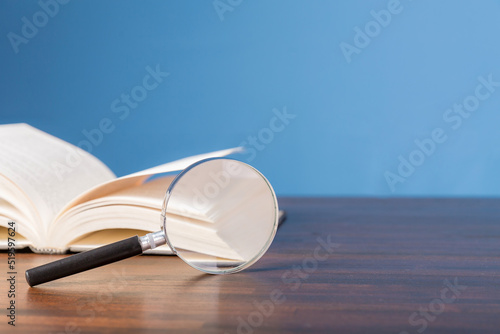 open book with magnifying glass on wooden desk in information library of school or university, concept for education,reading , study, copy space and blue background.