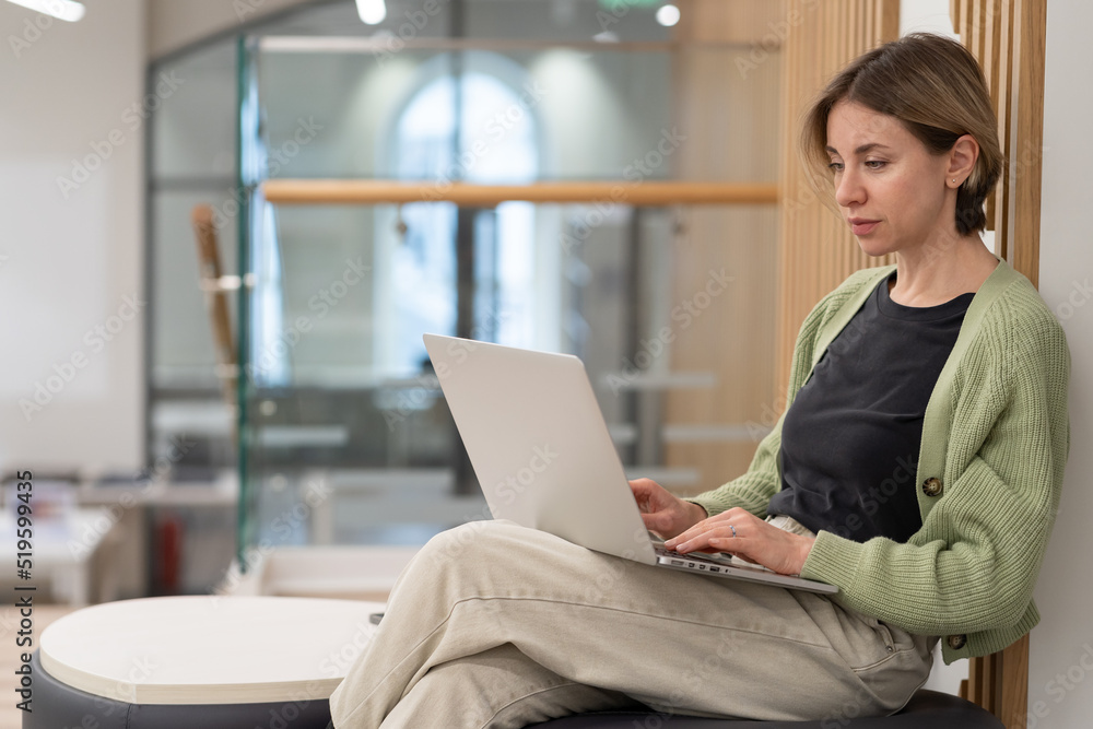 Focused pensive middle-aged woman sitting on ottoman with laptop, scandinavian lady freelance writer working remotely in coworking space, serious concentrated mature female remote worker in library Focused pensive middle-aged woman sitting on ottoman with laptop, scandinavian lady freelance writer working remotely in coworking space, serious concentrated mature female remote worker in library