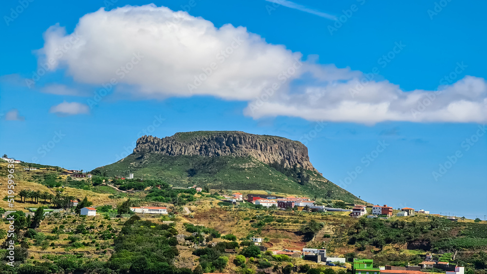 Panoramic view on massive volcanic plug Fortaleza de Chipude overlooking western coast of La Gomera, Canary Islands, Spain, Europe. Village in valley embedded lush green hills. Road to Valle Gran Rey