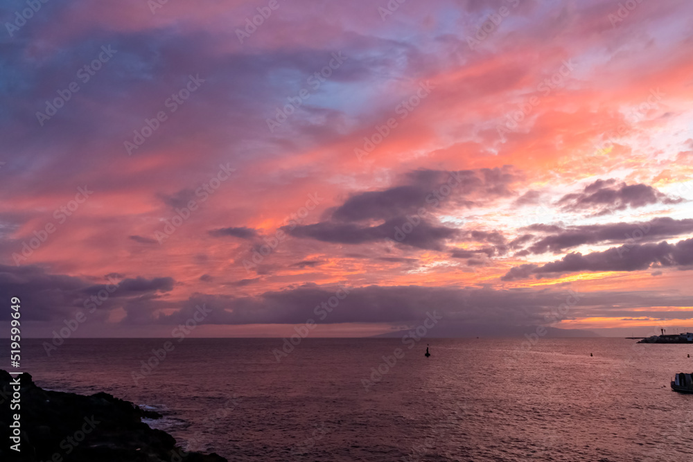 Obraz premium Romantic sunset over the sea in summer seen from lookout Cypelek Los Cristianos, Tenerife, Canary Islands, Spain, Europe. Vibrant colours of the clouds. Vacation vibes on the Atlantic Ocean. Awe