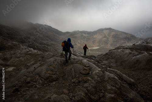 two men hiking toward a mountain hut in overcast bad weather amidst a barren mountain landscape with dramatic lighting conditions