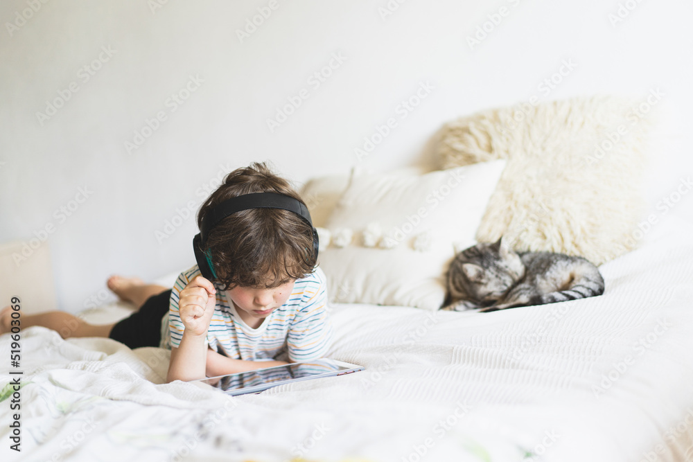 Cute boy lying on the bed at home and playing with pc tablet or reading online and listening to music with wireless headphones Cute boy lying on the bed at home and playing with pc tablet or reading online and listening to music with wireless headphones