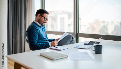 Confident entrepreneur analyzing documents while sitting at desk in office.