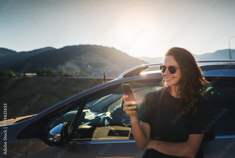 Young beautiful woman traveling by car in the mountains using smartphone at sunset, summer vacation and adventure