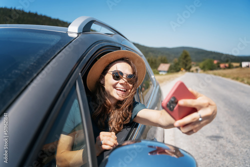 Happy young woman driver traveler in hat and sunglasses sitting behind the wheel of car making selfie using smartphone