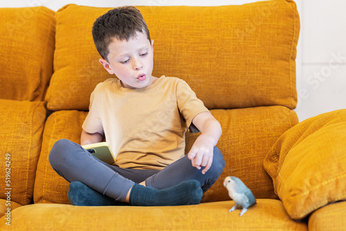 Boy sitting on a sofa whistling to a parakeet bird photo