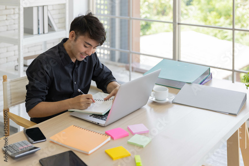 An Asian young business man work from home using computer and notebook together to manage his financial status