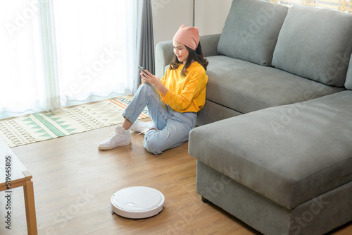 Young happy woman relaxing and using smartphone in living room while Robotic vacuum cleaner working © tonefotografia