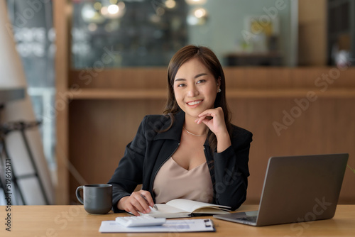 Smiling beautiful Asian businesswoman sitting at work looking at camera using laptop and computer while taking notes and working on documents at the office.