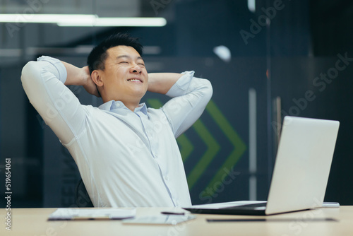 A break at work. A young Asian man is resting at the workplace at the desk in the office. He sits with his hands behind his head, closes his eyes, rests.