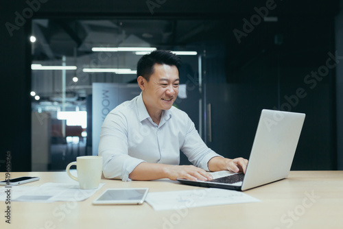 Young handsome male Asian freelancer conducts a video meeting on a video call from a laptop. Sitting at the table in a modern office, smiling, talking.