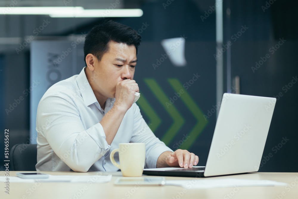 Sick at work. A young Asian businessman keeps coughing, covers his mouth at the desk in the office, feels bad, has an allergy, feels chest pain, needs help. Sick at work. A young Asian businessman keeps coughing, covers his mouth at the desk in the office, feels bad, has an allergy, feels chest pain, needs help.
