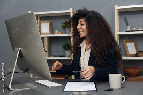 Cheerful enjoyed tanned adorable curly Latin businesswoman in jacket have video conferences with colleagues in office interior. Copy space Banner. Corporation leader lady uses modern computer in work