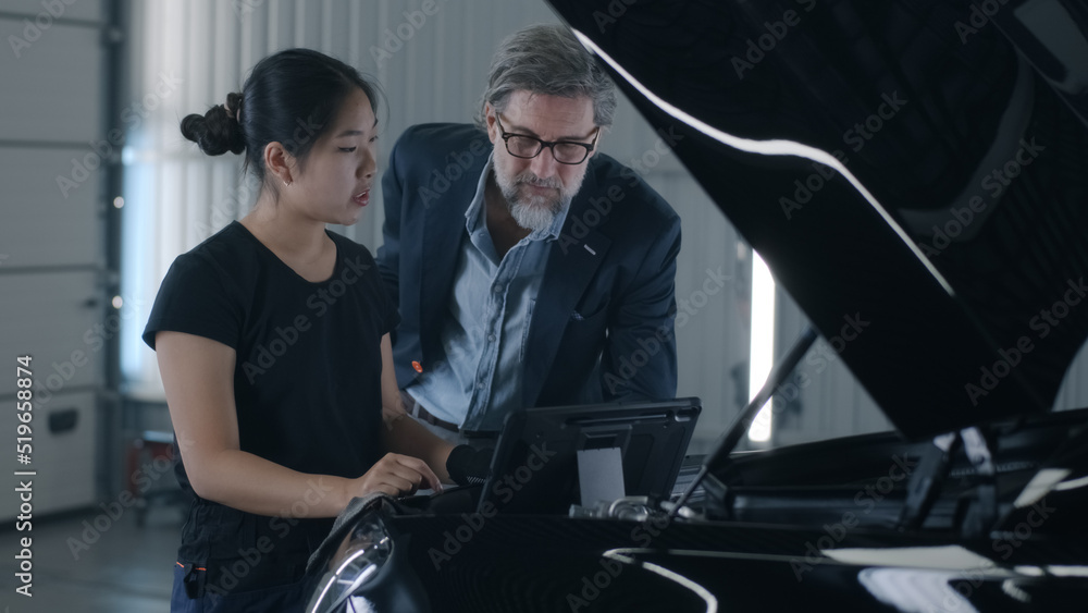 An asian woman mechanic consulting a man client in a car service, swiping on tablet and diagnosing a car with a breakdown An asian woman mechanic consulting a man client in a car service, swiping on tablet and diagnosing a car with a breakdown