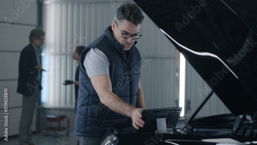 In a car service a male mechanic inspecting a car engine and making a diagnosis using a gadget while a female mechanic consulting a man