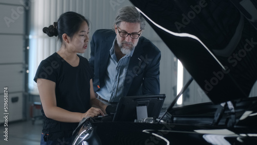 An asian woman mechanic consulting a man client in a car service, swiping on tablet and diagnosing a car with a breakdown