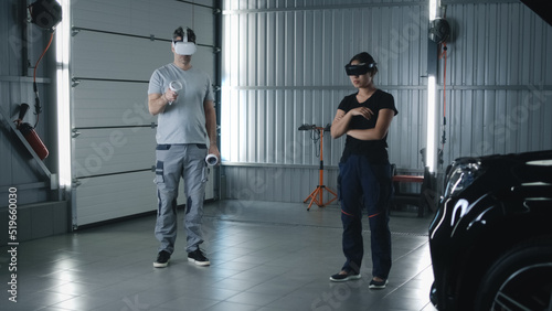 Man mechanic and female asian woman mechanic in virtual reality headsets with controllers talking while checking and diagnosing a car, with a malfunction in a car service
