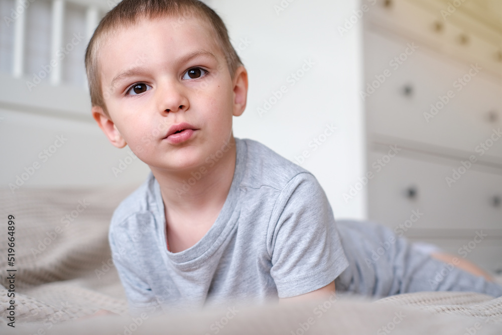 Close-up of a small happy caucasian boy in a gray t-shirt lies on the bed and smiles