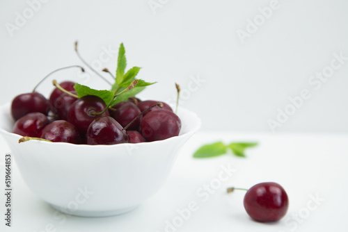 A white plate with a ripe burgundy cherry and a mint petal stands on a white background, there is a cherry and mint leaves next to it, a side view with a place for text. High quality photo © Ксения Мнасина