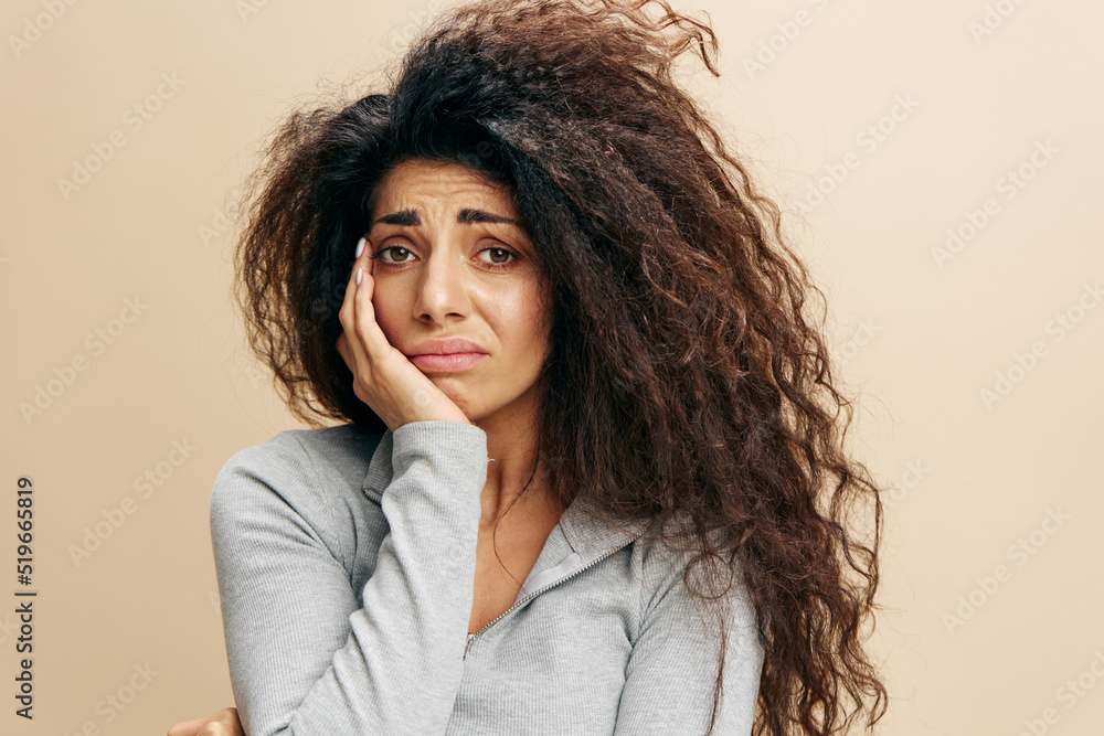 Young upset sad worried caucasian curly woman 20s wearing casual gray t-shirt look to you touch cheek isolated on plain pastel light beige background studio portrait. People lifestyle concept Young upset sad worried caucasian curly woman 20s wearing casual gray t-shirt look to you touch cheek isolated on plain pastel light beige background studio portrait. People lifestyle concept