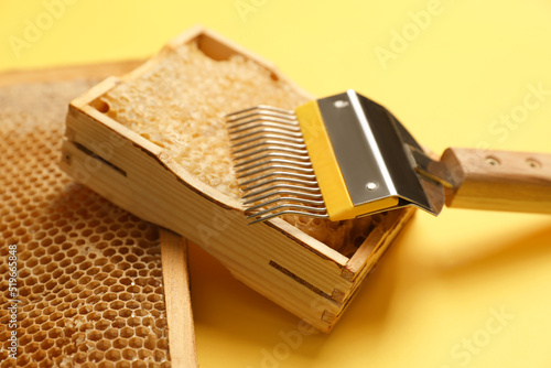 Hive frames with honeycombs and uncapping fork on yellow background, closeup. Beekeeping