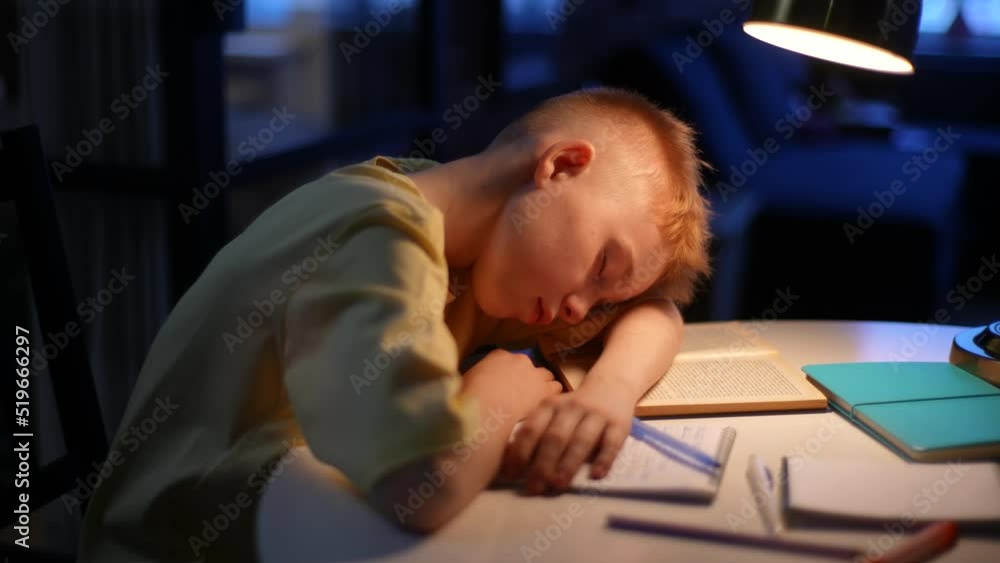 Tired little schoolboy sleeping at table with books and copybook under light of lamp in dark room. Concept of many homework and examinations stressful for young children. Shooting in slow motion. Tired little schoolboy sleeping at table with books and copybook under light of lamp in dark room. Concept of many homework and examinations stressful for young children. Shooting in slow motion.