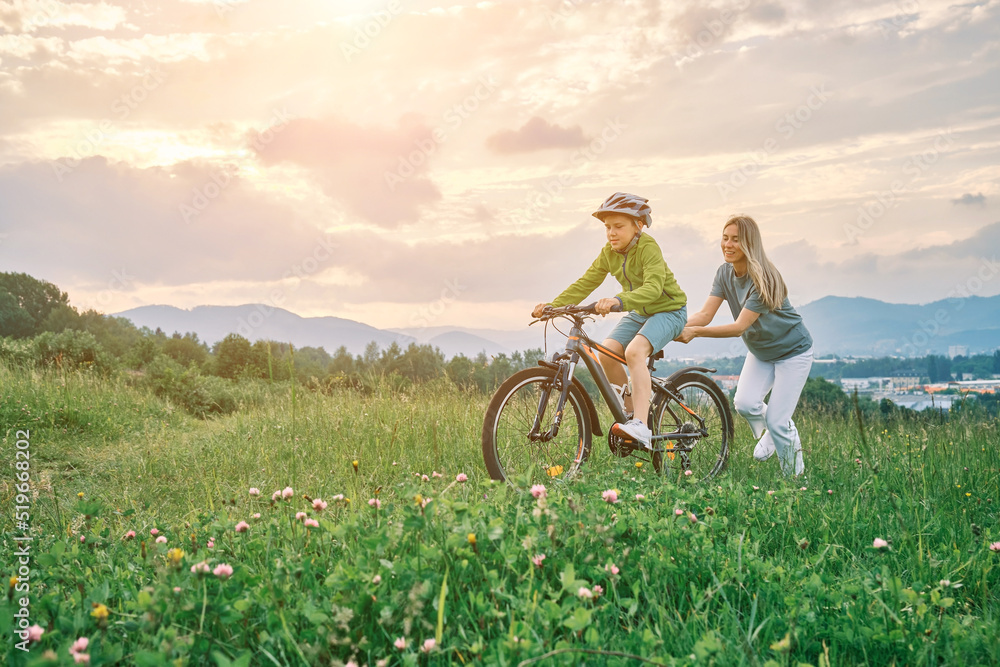 Obraz premium Mother teaching son to ride bicycle. Happy cute boy in helmet learn to riding a bike in park on green meadow in summer day at sunset time. Family weekend. 4K video Slow motion 