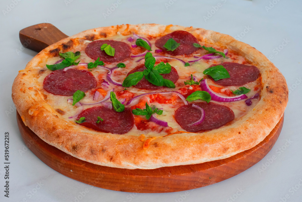 Delicious homemade pepperoni pizza with a thick crust on a wooden board with basil and red onions on a white marble kitchen top 