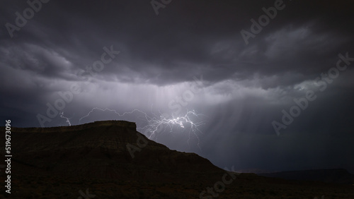 A bolt of lightning reveals heavy storm clouds and sheets of rain falling behind the silhouette of Smiths Mesa during a summer monsoon storm in Southern Utah. 