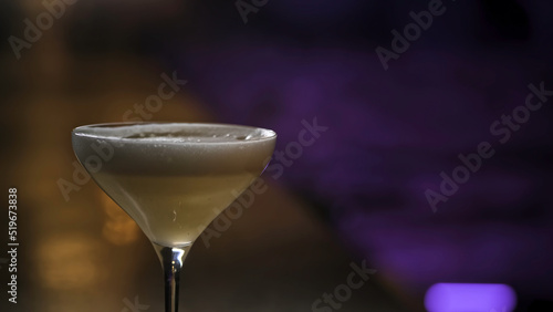 Close up of alcoholic drink with foam in martini glass. Stock footage. A cocktail of yellow color standing on purple and brown background of a dark room.