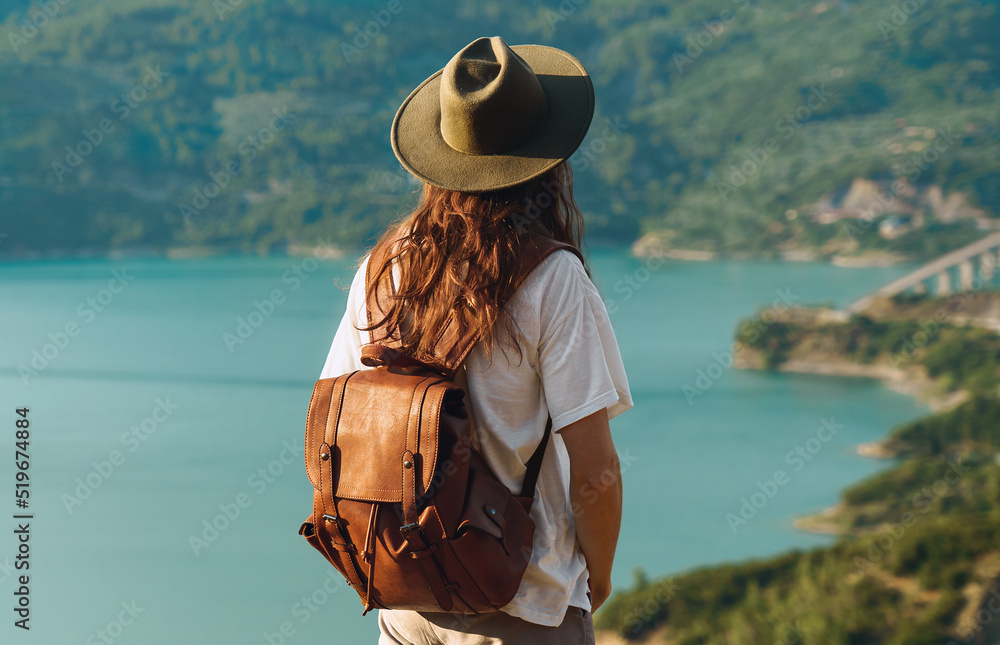 Back view of traveler man with hat and backpack looking at scenic view of lake and mountains from viewpoint.
