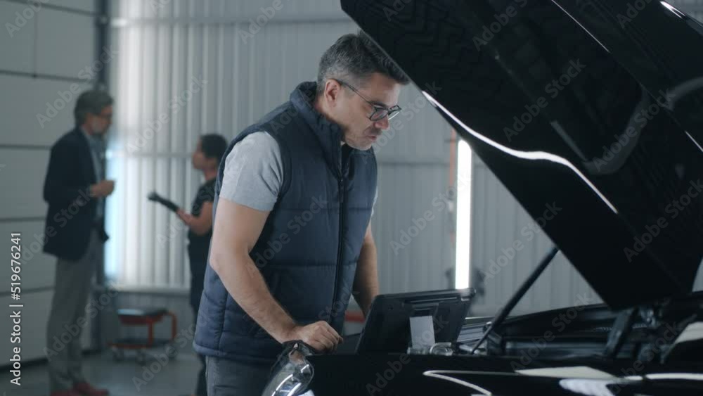 Handheld shot of a service manager worker or mechanic in repair shop inspecting a car engine and making a diagnosis using a gadget, while a female mechanic consulting a man