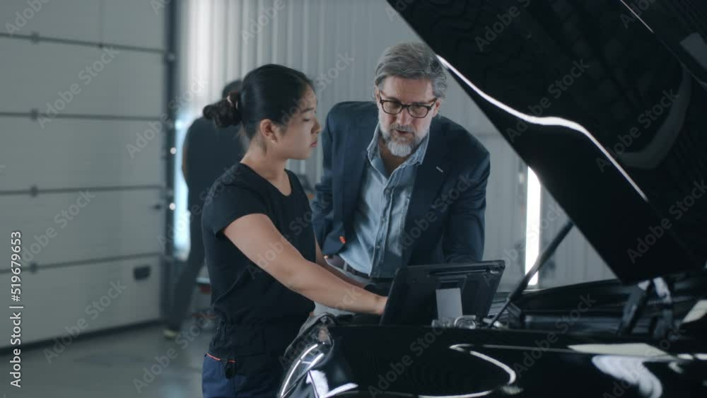 An asian female mechanic in repair shop consulting a male client in a car service, swiping on tablet and diagnosing a car with a breakdown An asian female mechanic in repair shop consulting a male client in a car service, swiping on tablet and diagnosing a car with a breakdown