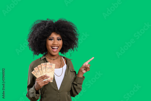 woman holding money in one hand, and point to empty space, afro woman holding brazilian money, isolated on green background photo