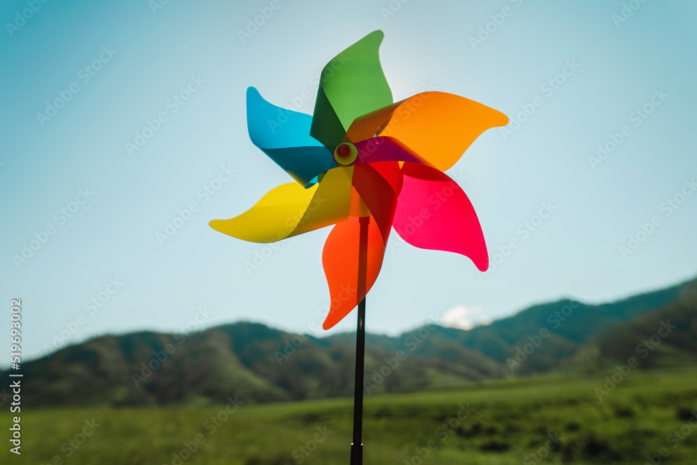 A multi-colored children's windmill, pinwheel in the background of sky and a green landscape. Clean green energy symbol A multi-colored children's windmill, pinwheel in the background of sky and a green landscape. Clean green energy symbol