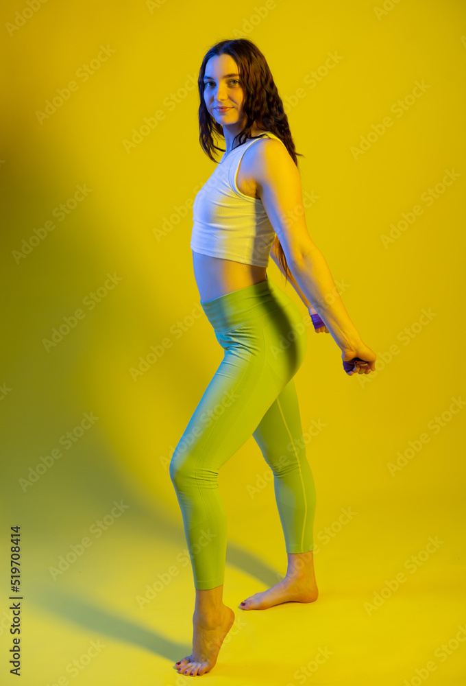 Athletic girl in green leggings and a blue t-shirt does exercises for the buttocks with a resistance band. Fitness woman exercising. yellow background.