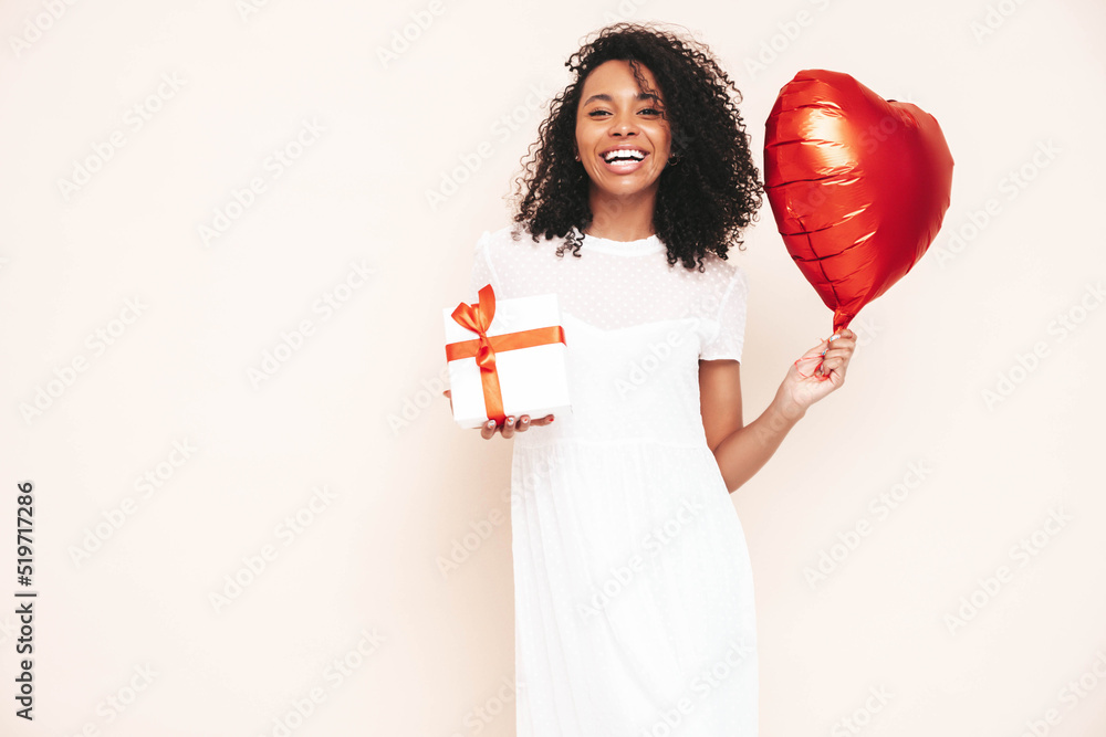 Beautiful black woman with afro curls hairstyle. Smiling model dressed in white summer dress. Sexy carefree female posing near blue wall in studio. Holding heart air balloon and gift box. Isolated Beautiful black woman with afro curls hairstyle. Smiling model dressed in white summer dress. Sexy carefree female posing near blue wall in studio. Holding heart air balloon and gift box. Isolated